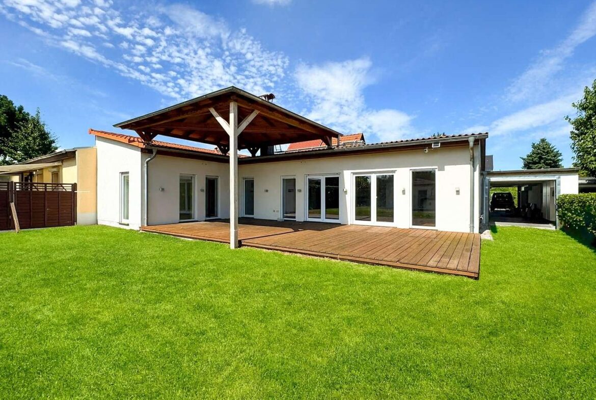 Backyard view of a modern single-story white house with a large wooden deck under a shaded pergola and a green lawn under a bright blue sky.