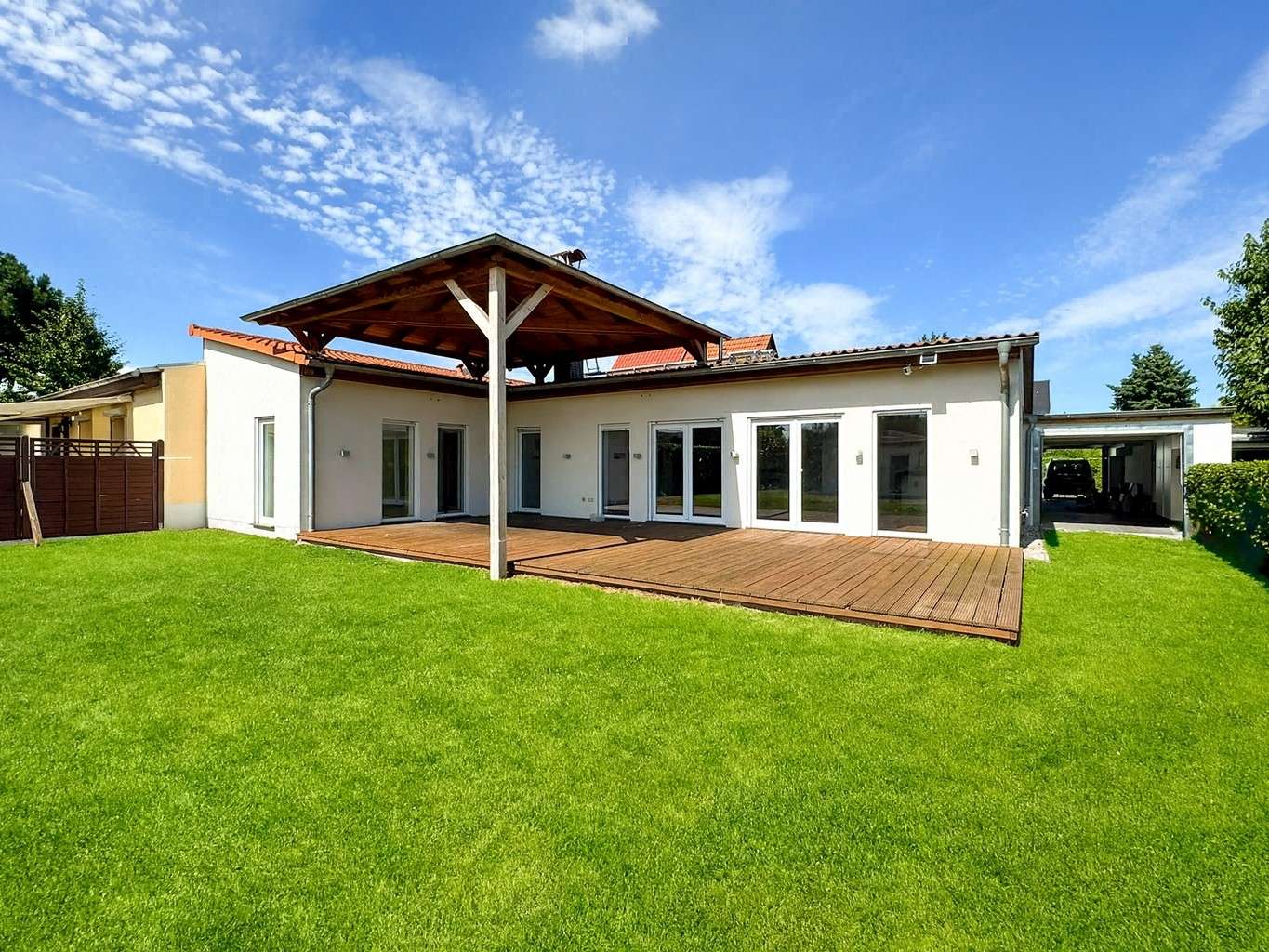 Backyard view of a modern single-story white house with a large wooden deck under a shaded pergola and a green lawn under a bright blue sky.