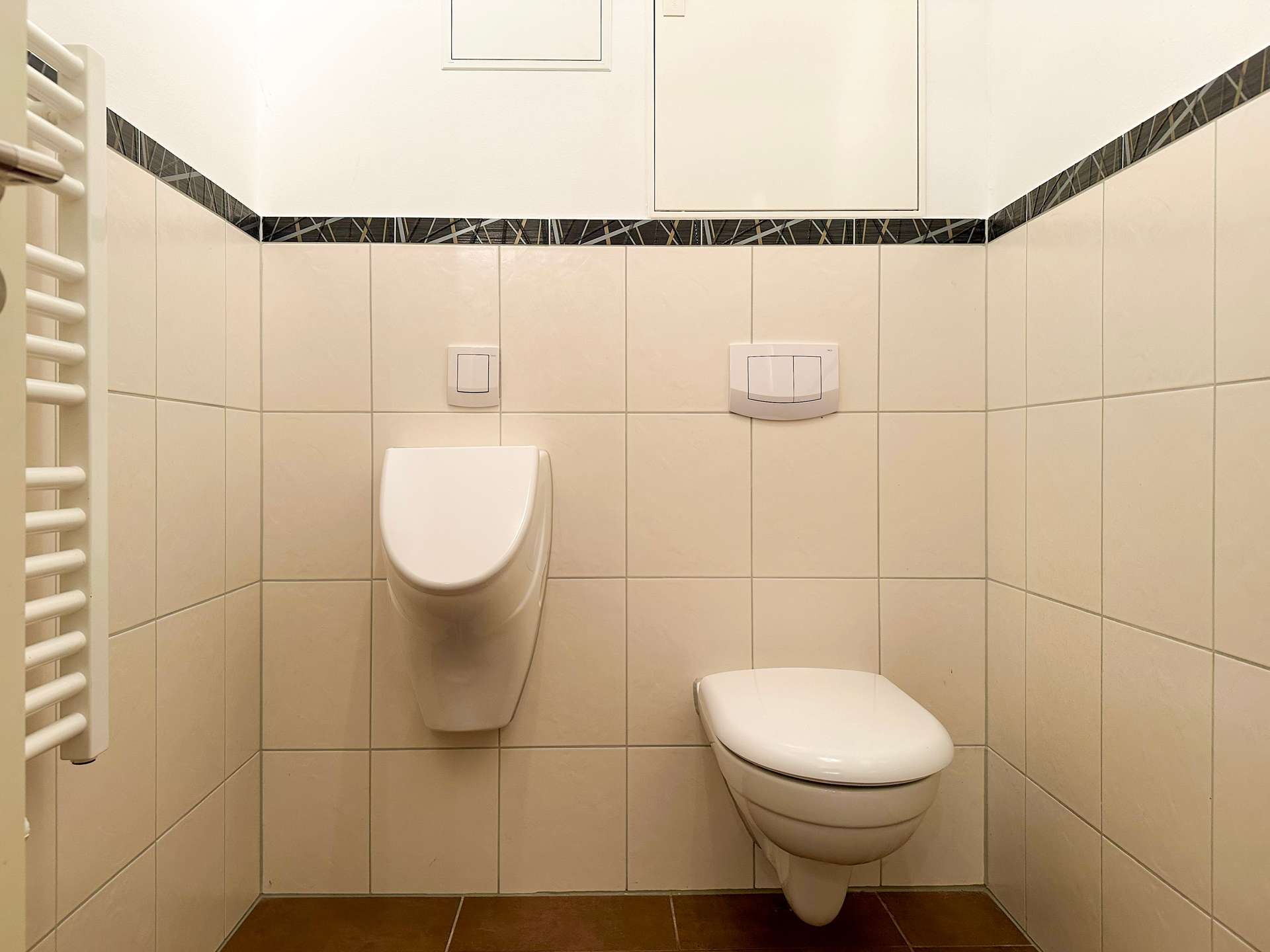 Small bathroom with a wall-hung toilet, dual-flush panel on the wall, beige tiled walls, and a dark decorative tile border near the top left-to-right.