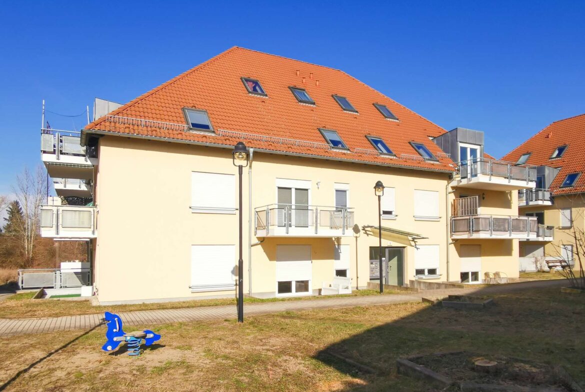 Apartment building with a red-tiled sloped roof and balconies under a bright blue sky; a blue children's playground rocker in the foreground.