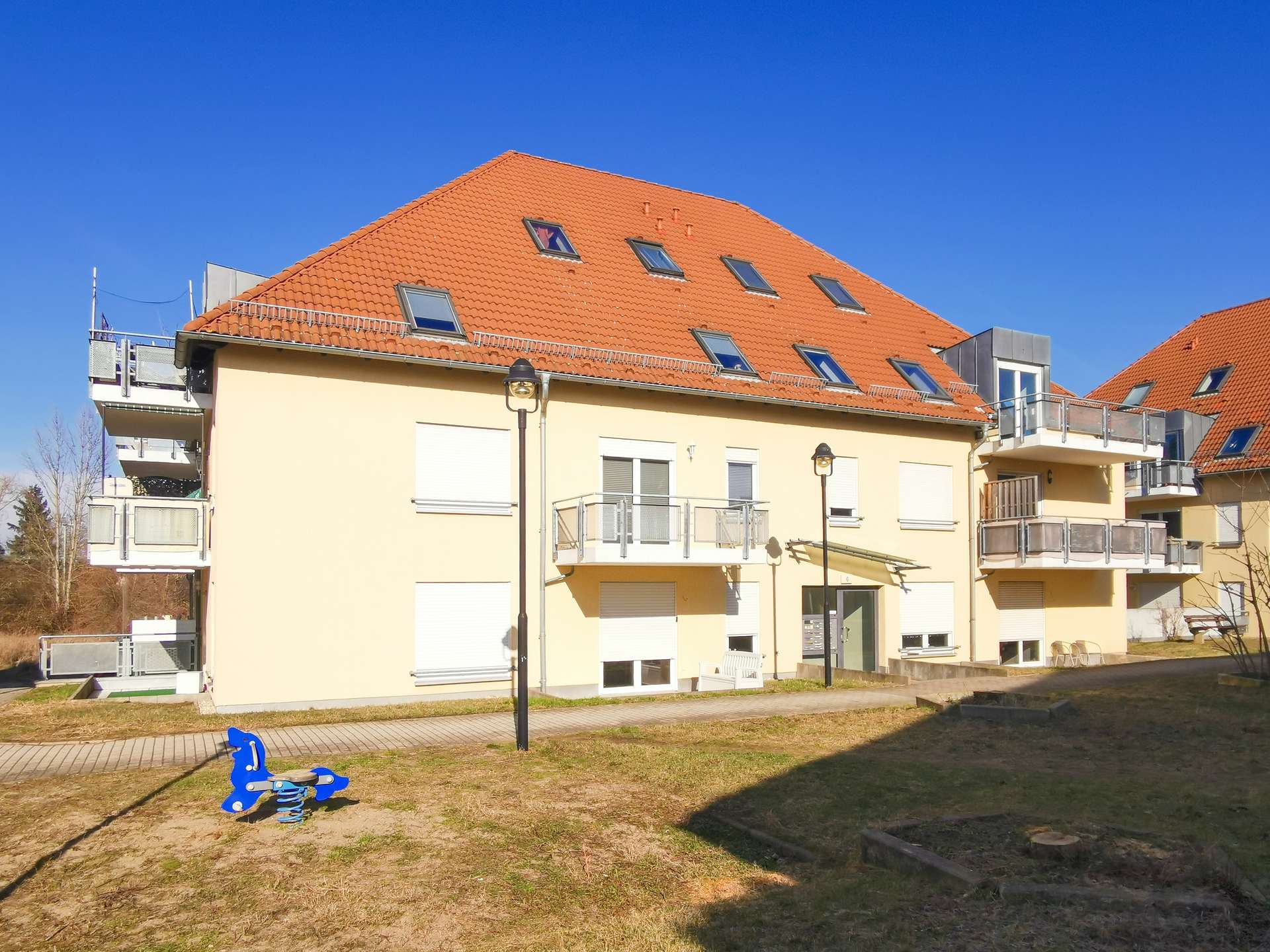 Apartment building with a red-tiled sloped roof and balconies under a bright blue sky; a blue children's playground rocker in the foreground.