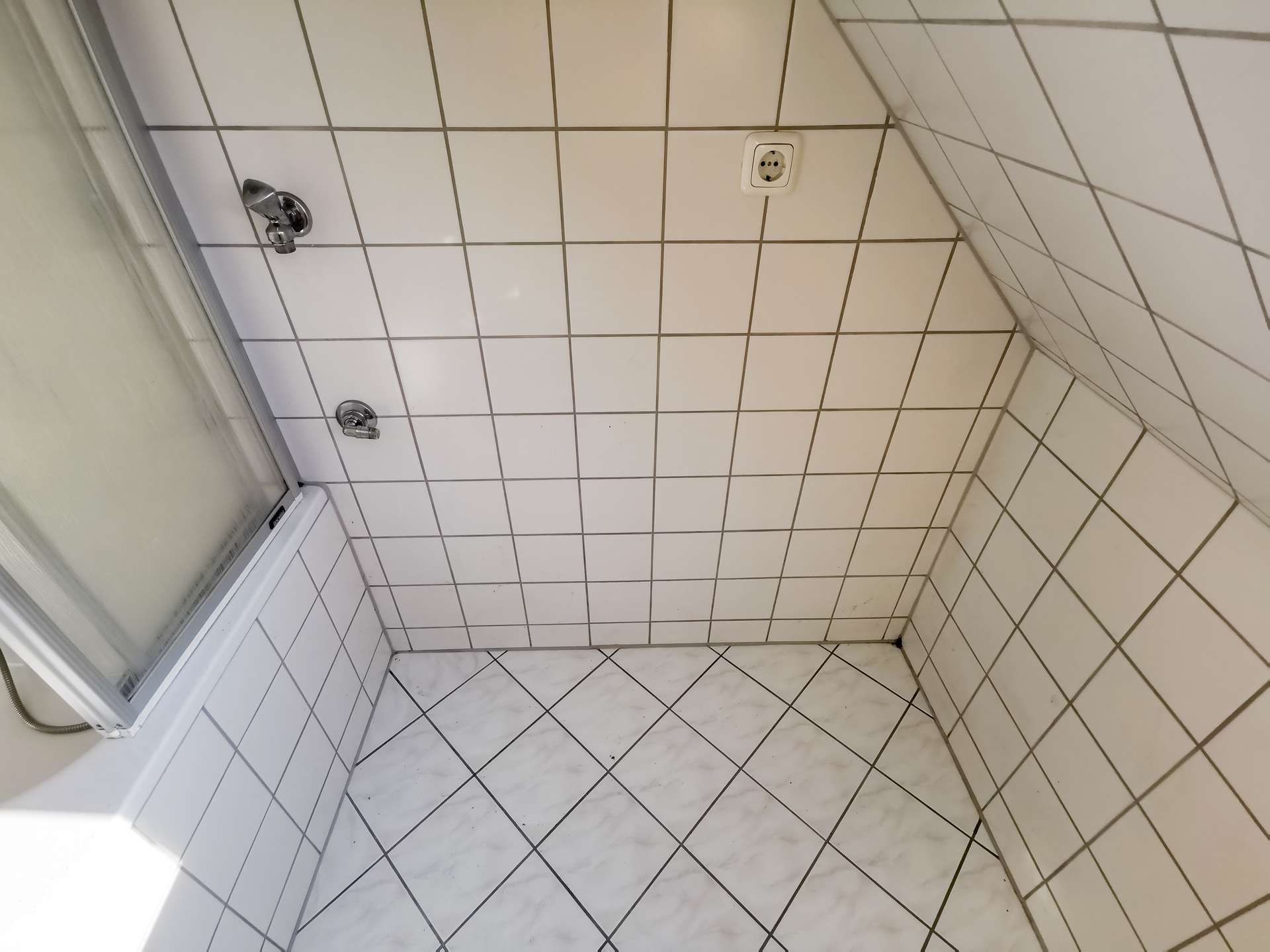 Shower stall with white square tiles, a glass door on the left, and two metal faucet handles on the wall opposite. A small electrical outlet is visible on the tiled wall.
