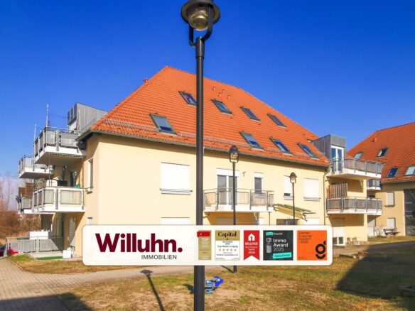 Large white Willuhn Immobilien sign with award badges in front of a beige apartment building with an orange tiled roof and blue sky.