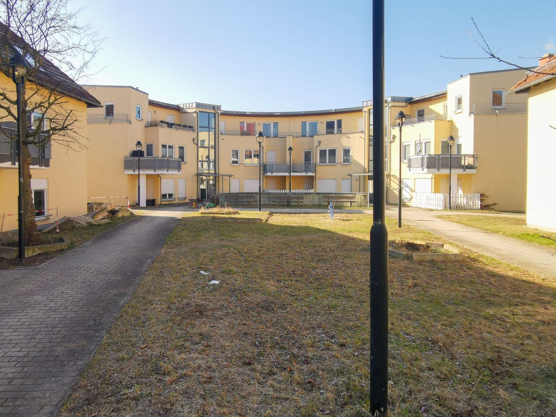 Courtyard of a yellow apartment complex with balconies, curved building, and a central black lamppost against a blue sky