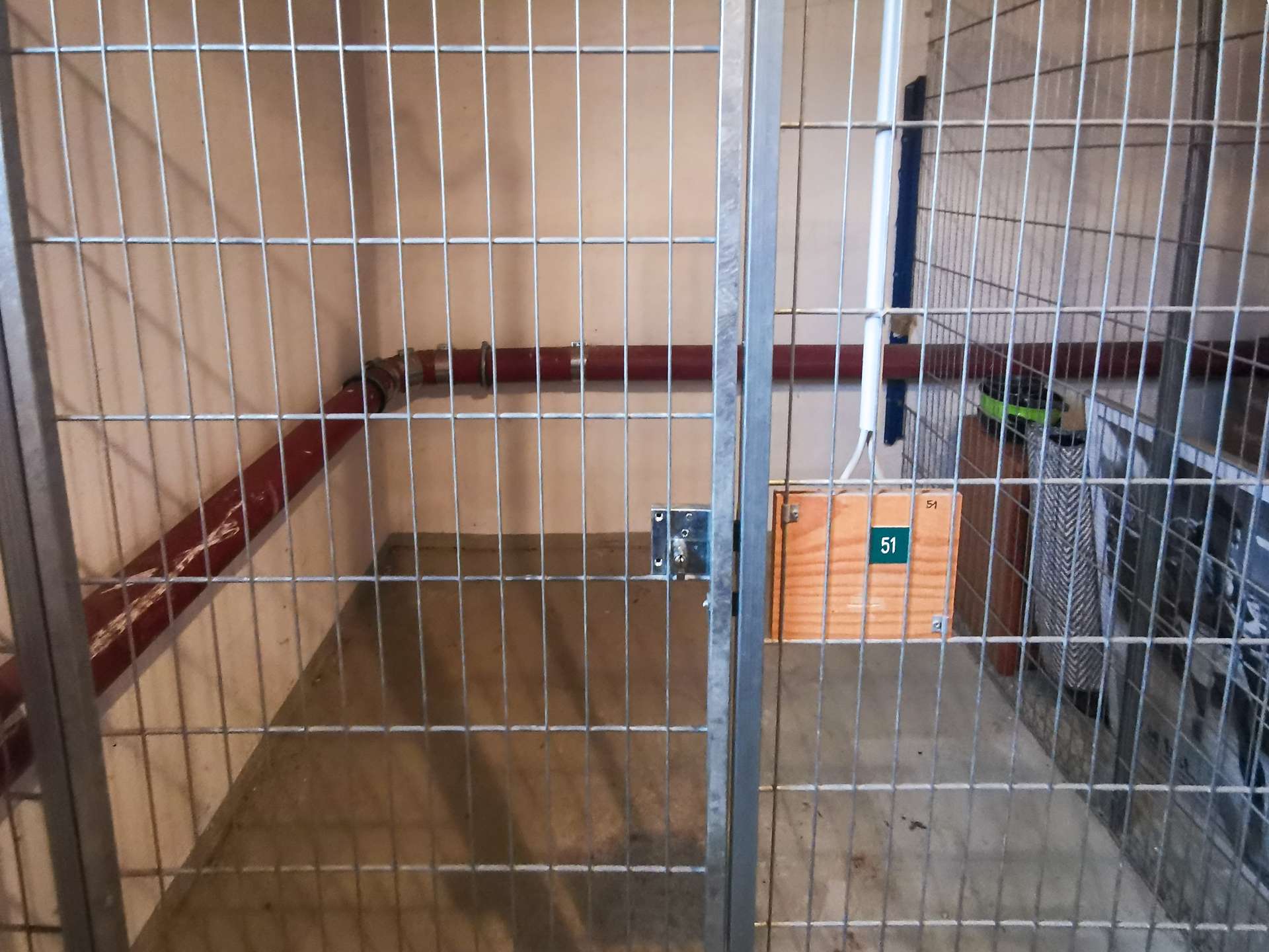 Empty metal cage with vertical bars; red pipes run along the back wall and a cardboard box labeled '51' sits in the corner.
