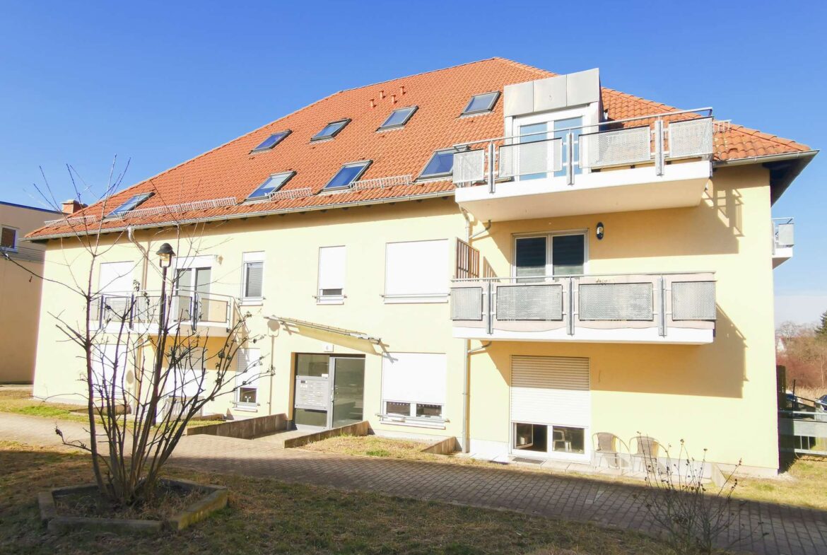 Apartment building with a red-tiled sloped roof, skylights, and balconies along a pale yellow facade under a blue sky.