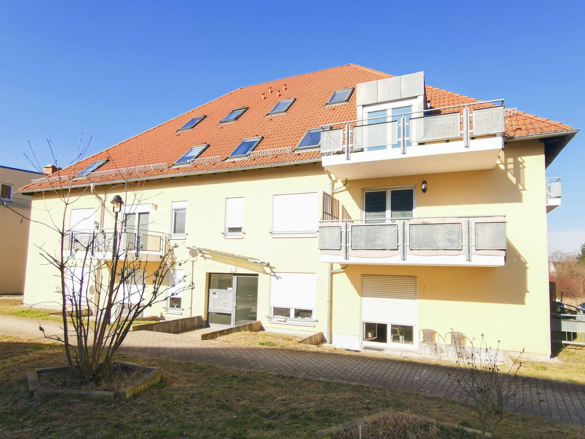 Apartment building with a red-tiled sloped roof, skylights, and balconies along a pale yellow facade under a blue sky.