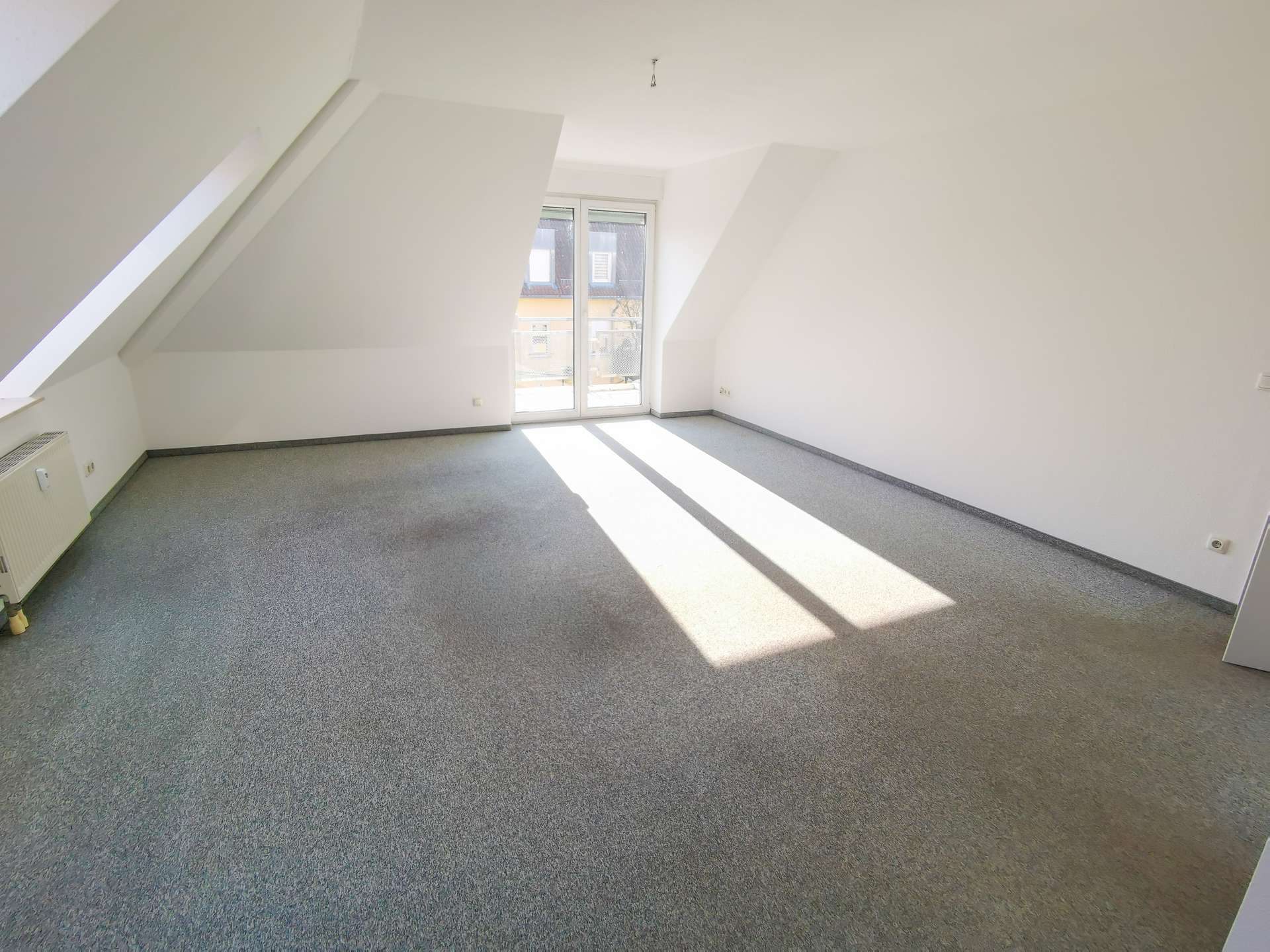 Bright empty attic room with slanted walls and a balcony door; sunlight casts long shadows on grey carpeted floor.