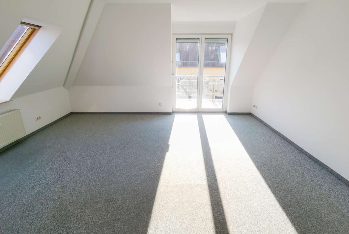 Bright empty attic room with slanted walls, large glass door to a balcony, and sunlight streaming across a gray carpet.