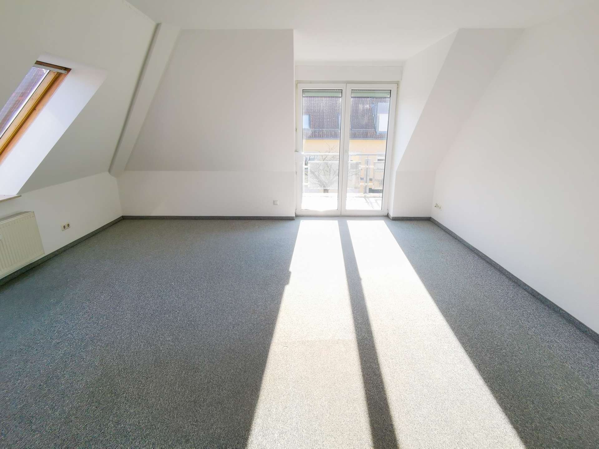 Bright empty attic room with slanted walls, large glass door to a balcony, and sunlight streaming across a gray carpet.