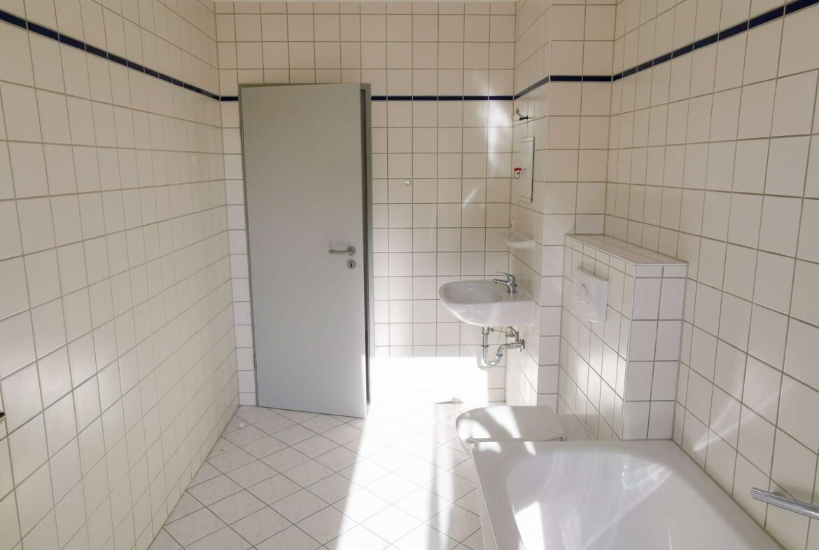 Bathroom with white square tiles and a dark blue accent line, gray door ajar, sink, mirror, and toilet visible.