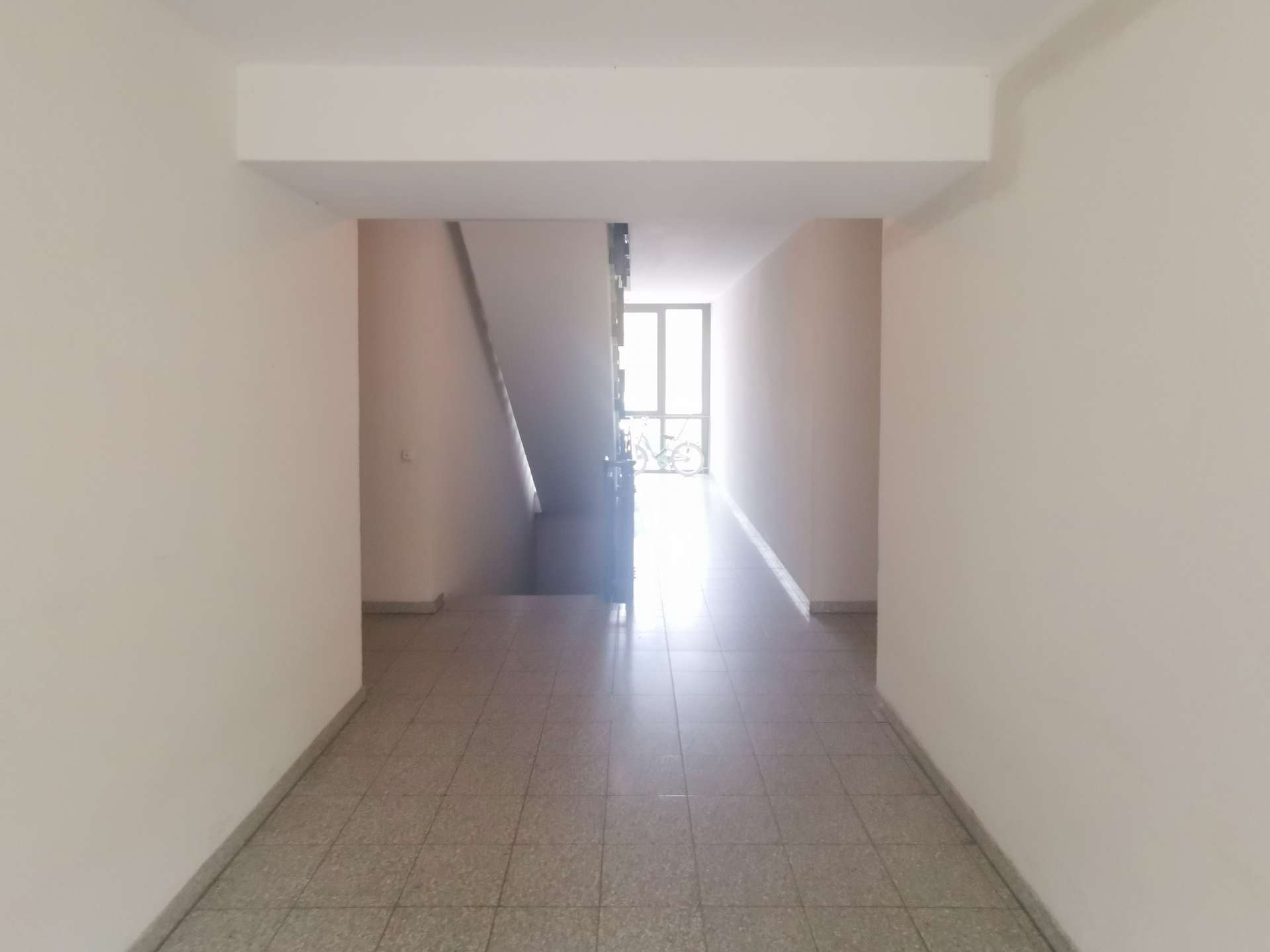 Long indoor hallway with stairs on the left, tile floor, and bright light at the far end where a bicycle is visible by the window railings.