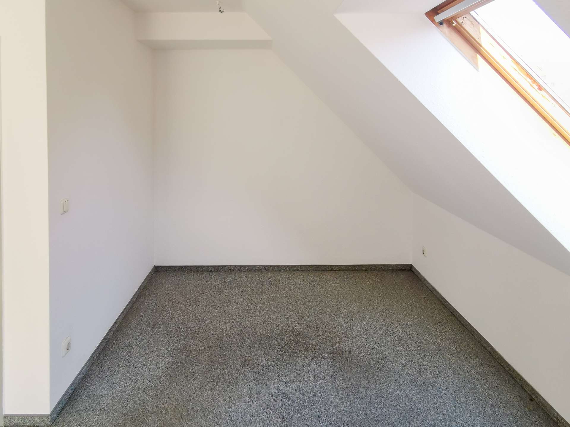 Empty attic-like room with white walls, a sloped ceiling, and a skylight on the right; gray speckled floor.