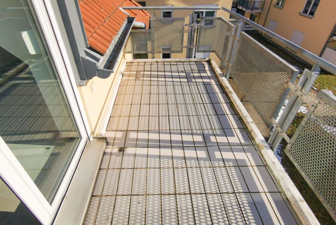 Small balcony with a metal grid floor and perforated railing, overlooking orange-tiled rooftops and a yellow apartment building across.
