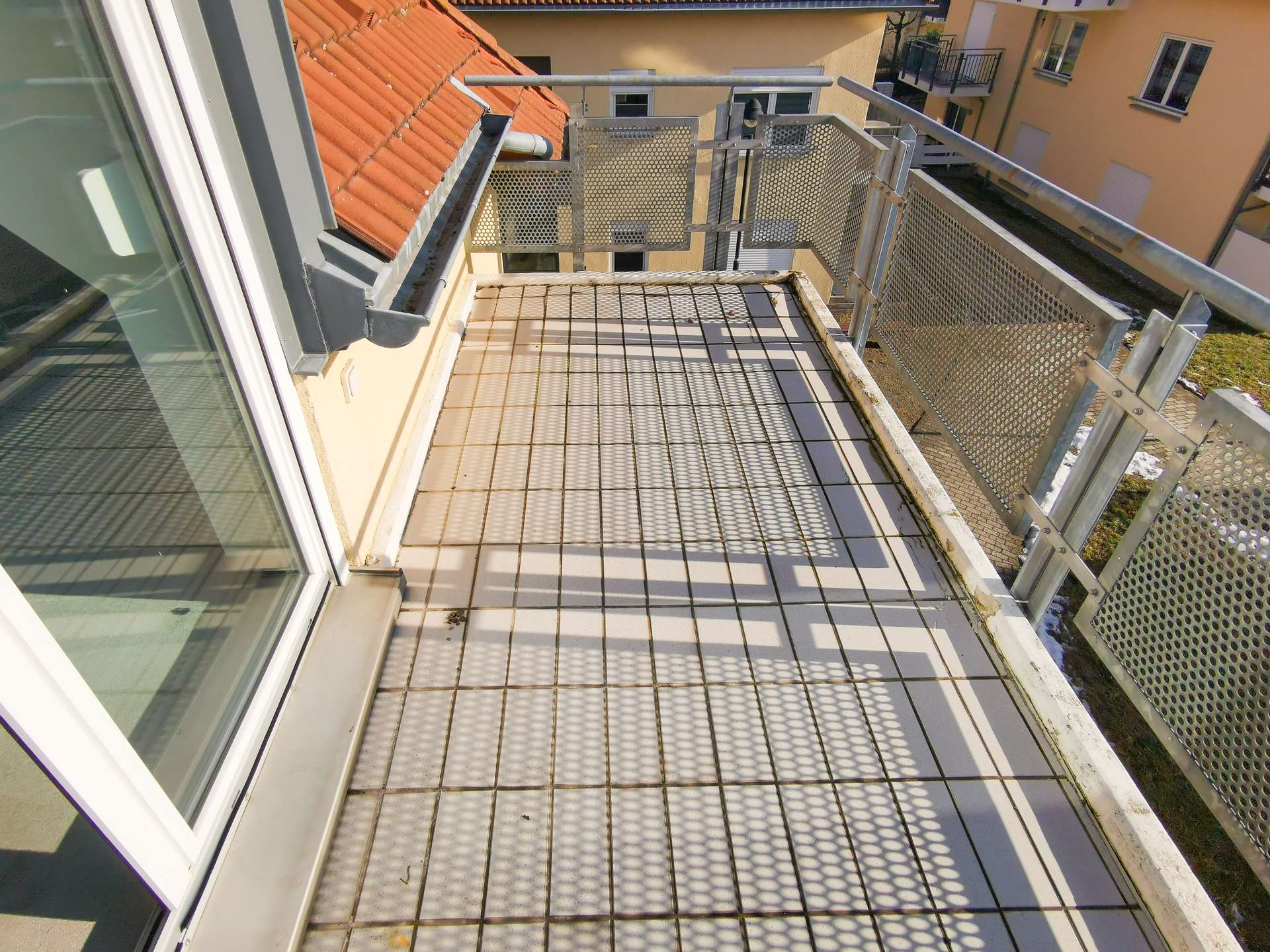 Small balcony with a metal grid floor and perforated railing, overlooking orange-tiled rooftops and a yellow apartment building across.