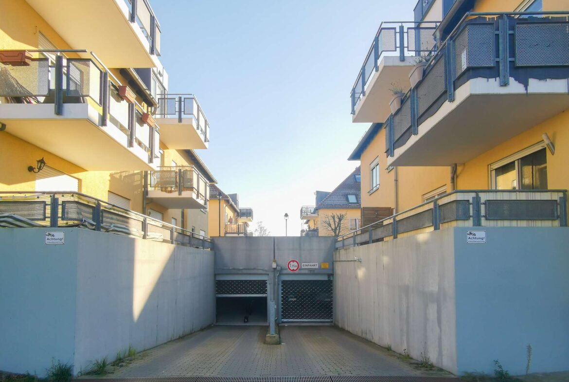 View down a narrow driveway between yellow apartment buildings with blue metal railings and balconies; gate at the far end and clear blue sky overhead.