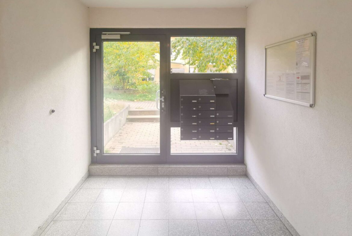 Entry hall with a glass-framed door and a row of black mailboxes; a notice board is on the right wall, with greenery visible outside.