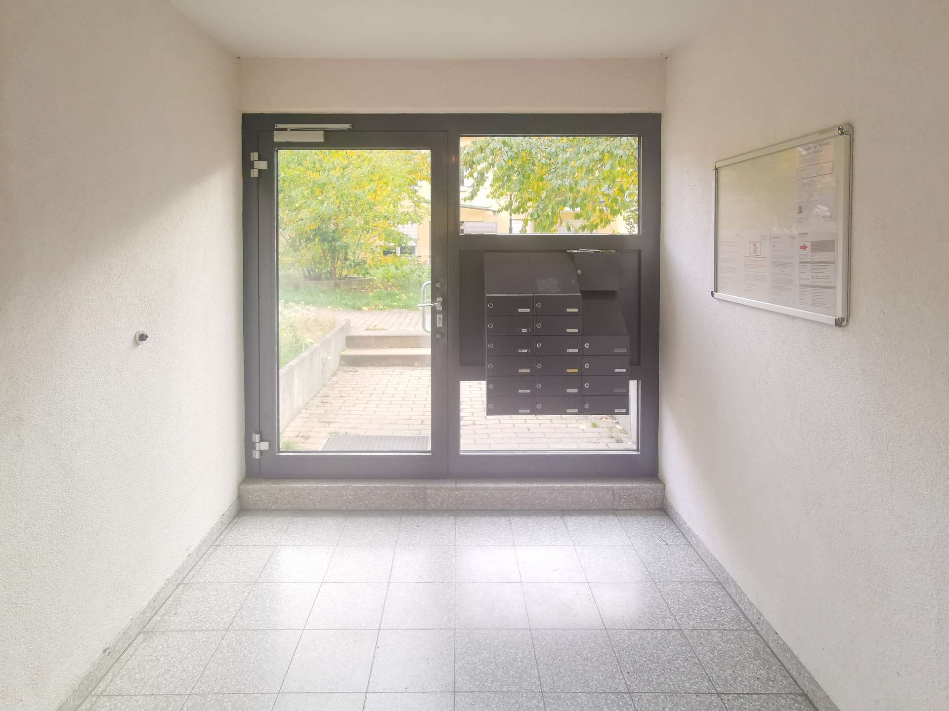 Entry hall with a glass-framed door and a row of black mailboxes; a notice board is on the right wall, with greenery visible outside.