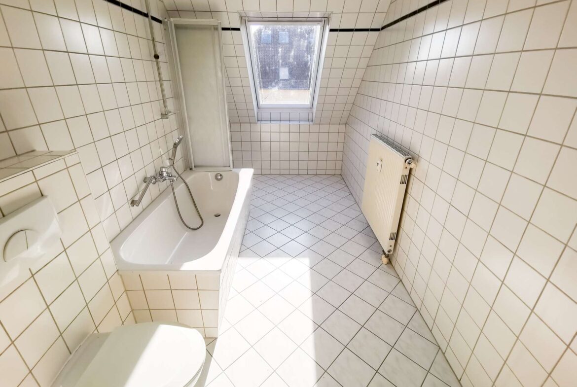 Small, narrow tiled bathroom featuring a white bathtub with a handheld shower near the left wall, a glass shower cubicle, a centered window, and a wall-mounted radiator on the right.
