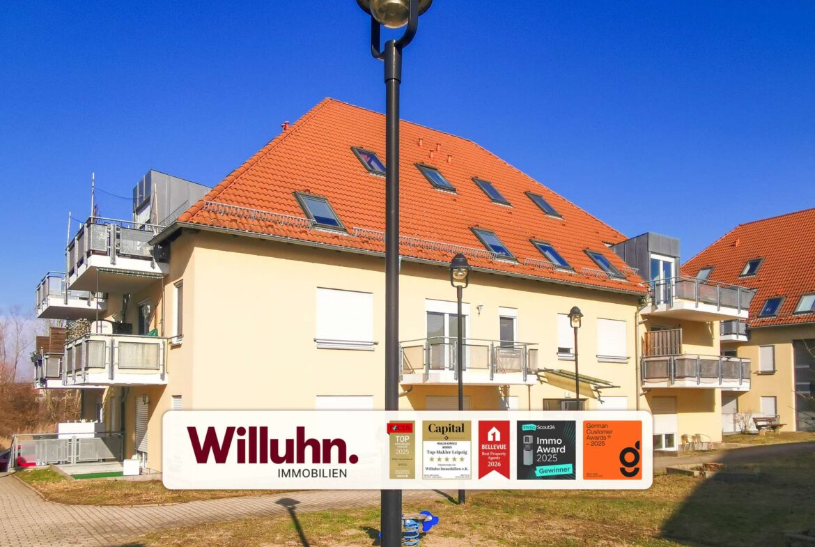 Residential building with a red tiled roof and balconies under a clear blue sky; a tall street lamp stands in front and a large Willuhn Immobilien sign with award badges is visible in the foreground.