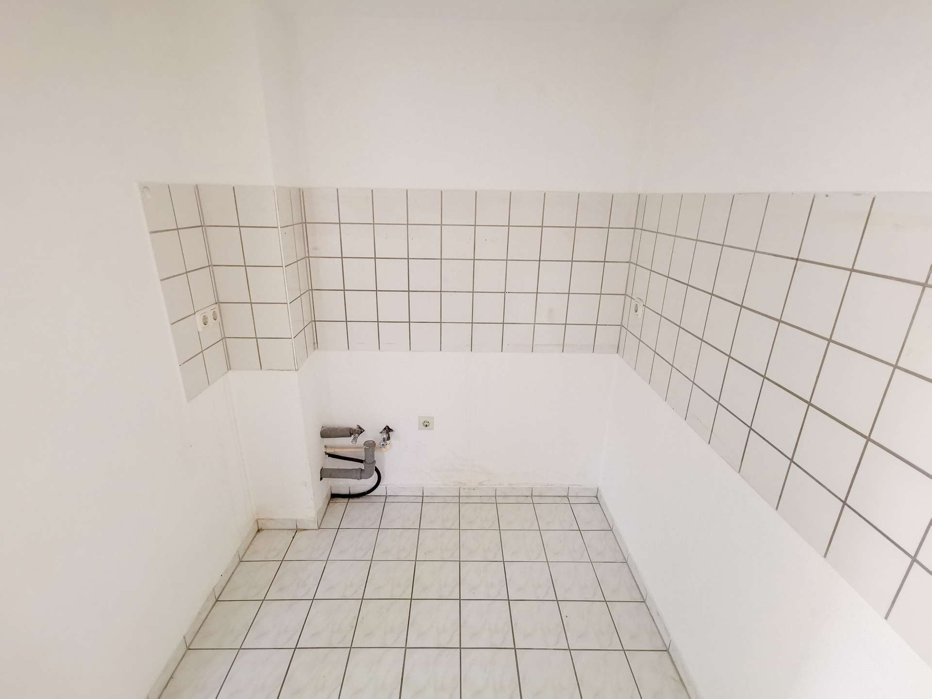 Bare utility corner with white tiled walls, exposed pipes and plumbing under a missing sink area.