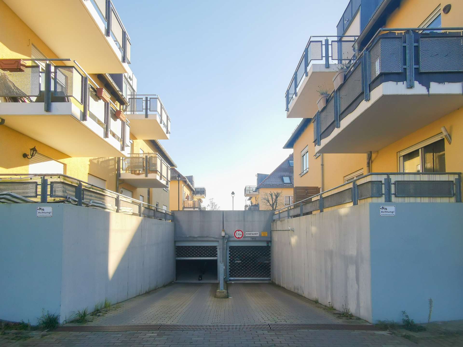 Underground parking entrance between yellow apartment buildings with blue railings and a metal gate at the end of a paved driveway.