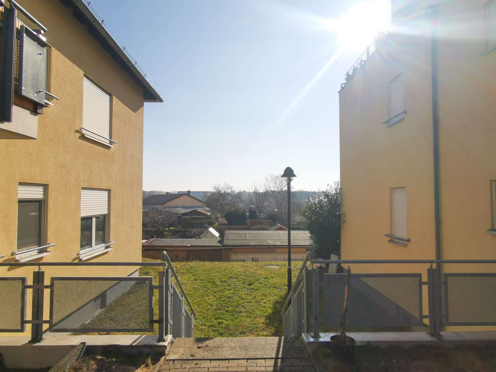 Sunny courtyard between two yellow apartment buildings, metal railings, steps, and a grassy area beyond; sun glare in the upper right.