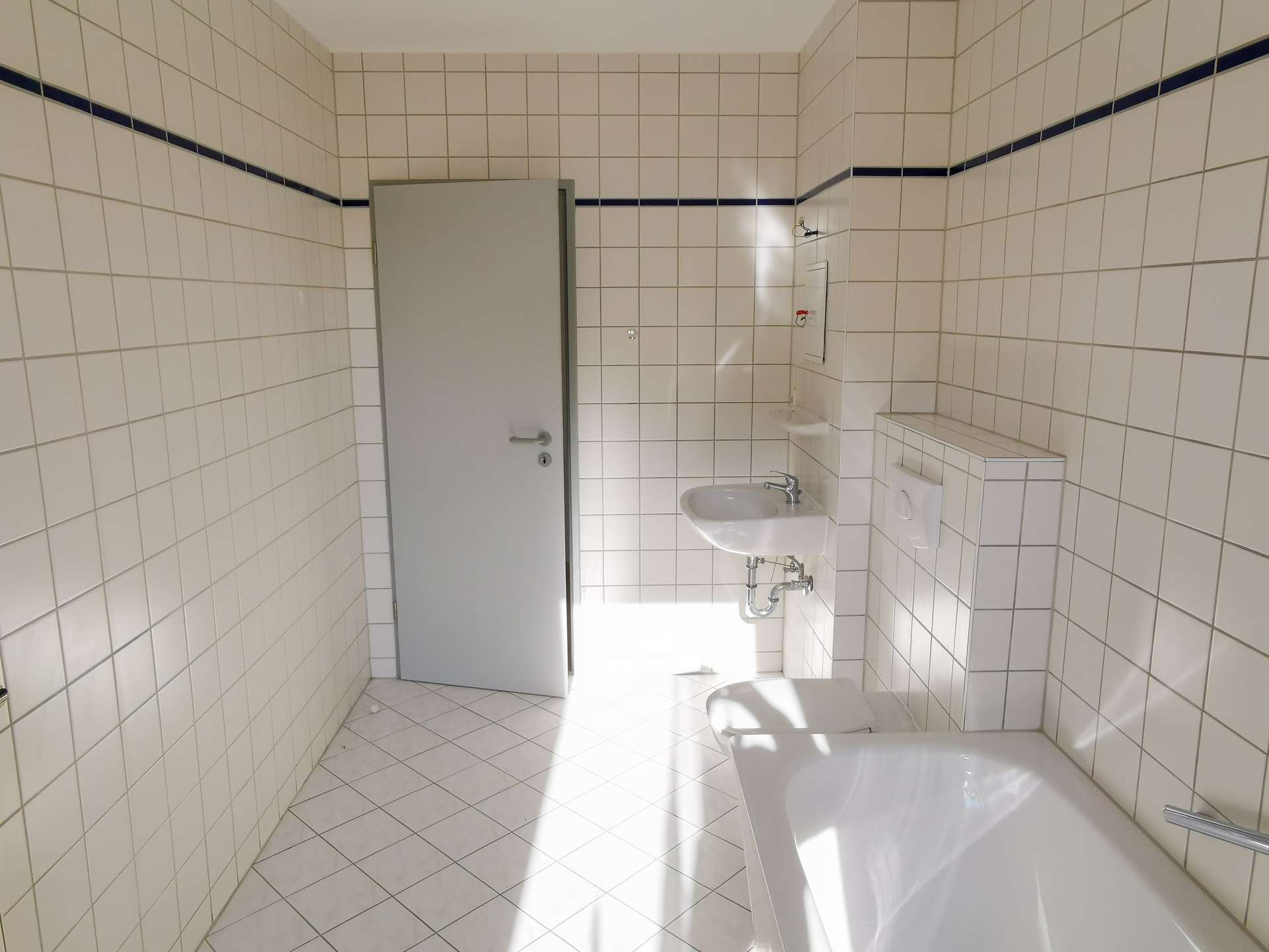 White tiled bathroom with a gray door, pedestal sink, and toilet along tiled walls, bright with natural light.