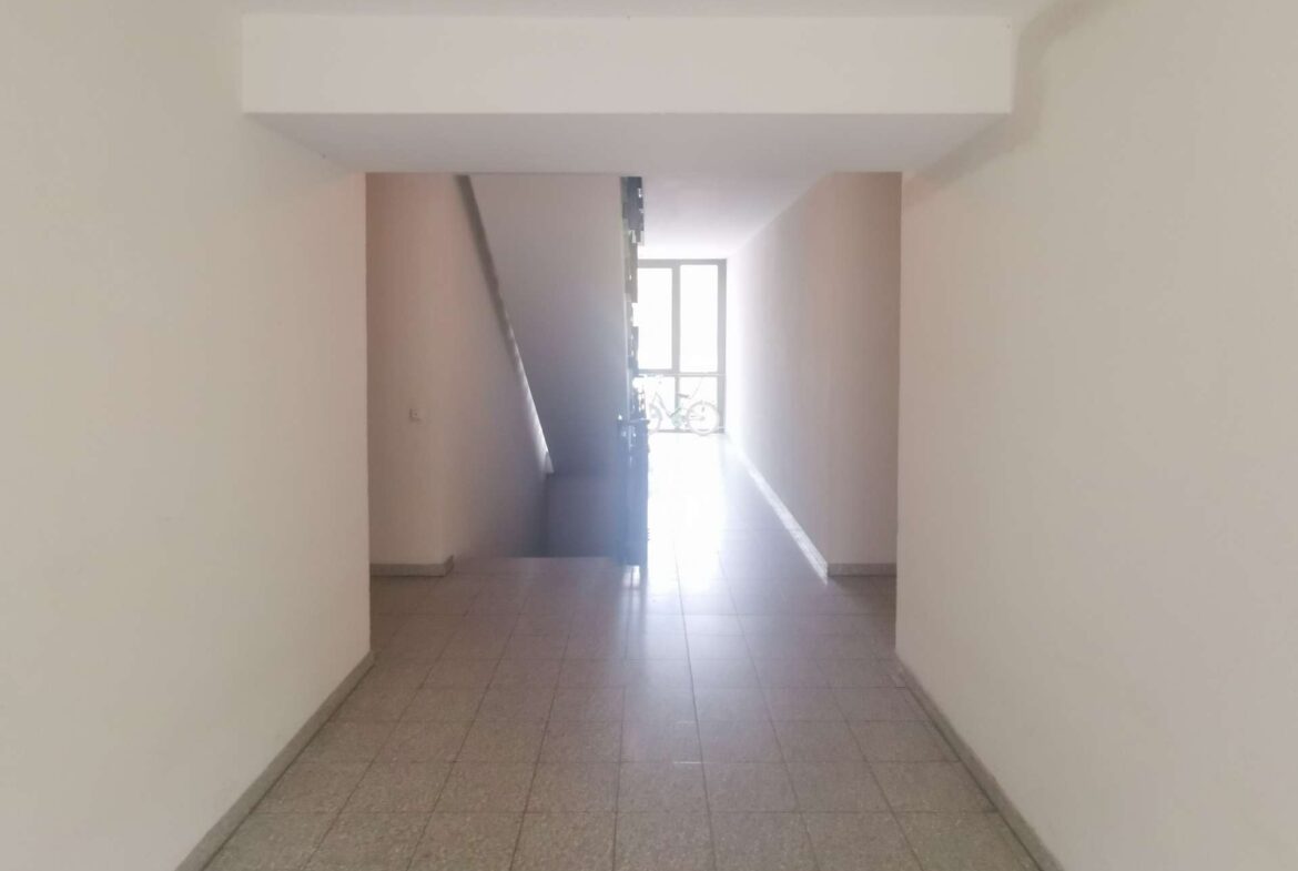 Indoor stairwell hallway with stairs on the left, beige walls, and a sunlit doorway at the far end where a bicycle is visible outside the window.