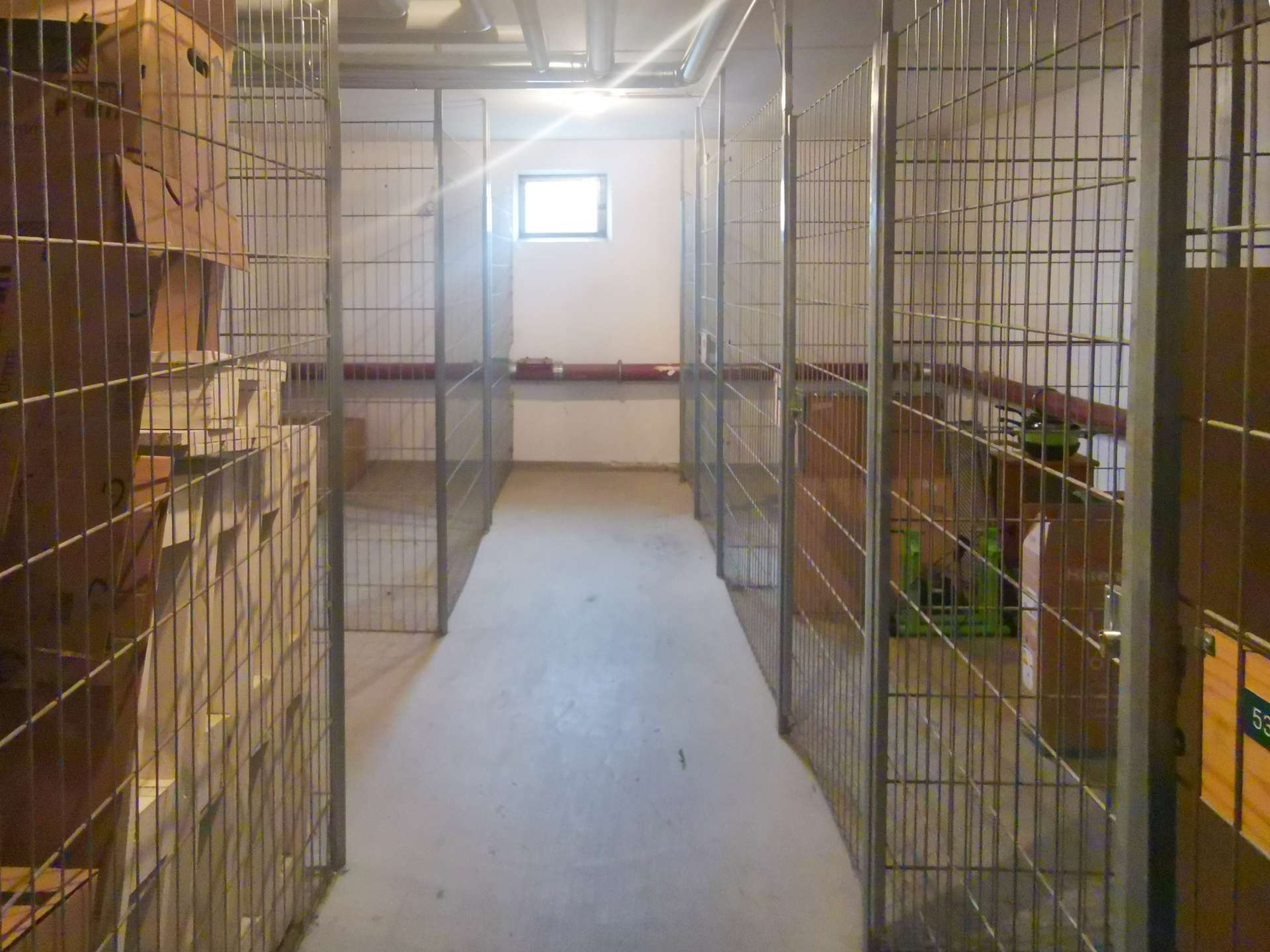 Wide hallway flanked by metal storage cages filled with boxes and shelves; a small window at the end lets in light.