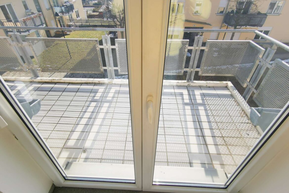 View from inside a room looking out through a glass door onto a metal balcony with perforated panels and tiled flooring.