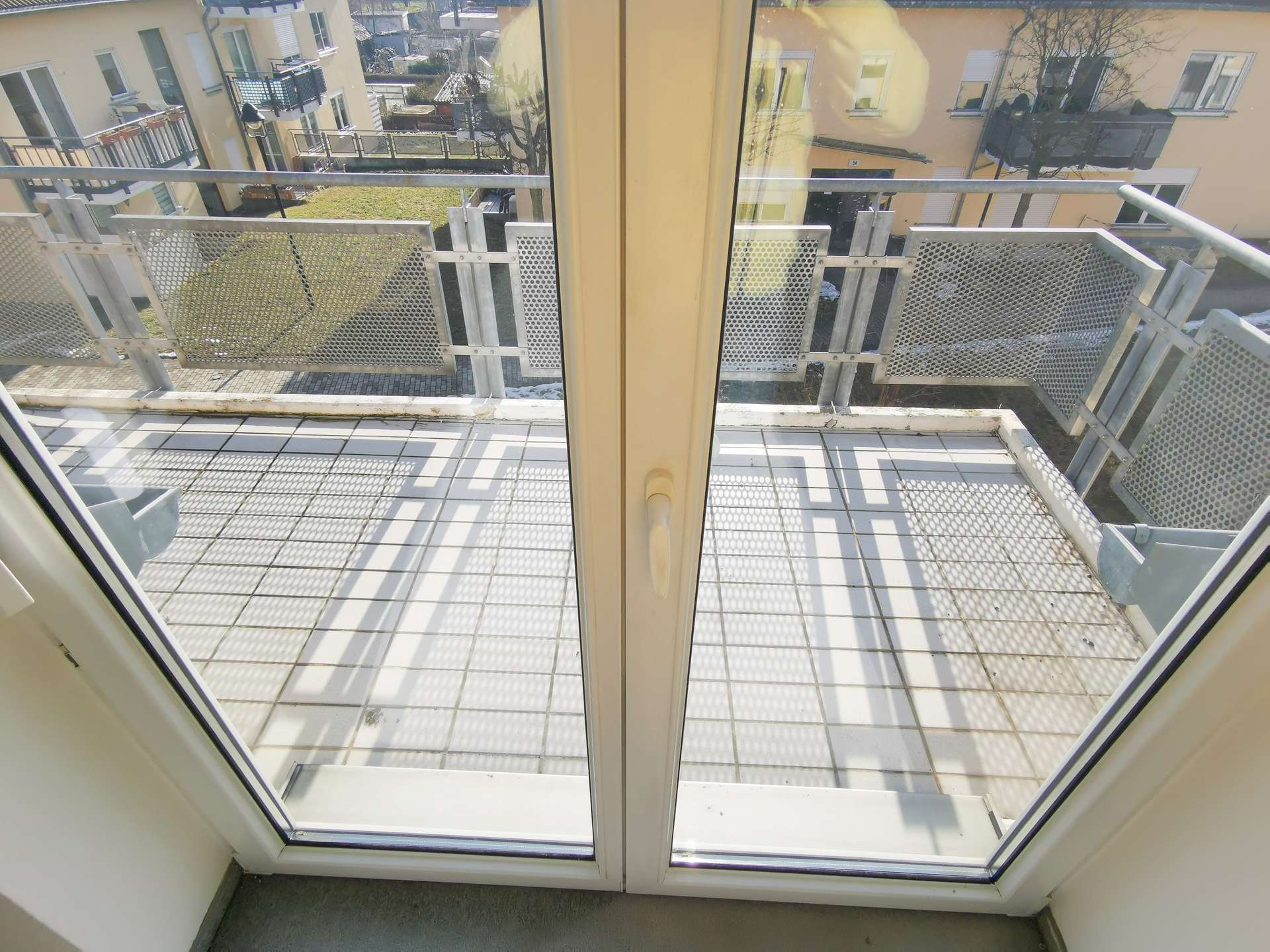 View from inside a room looking out through a glass door onto a metal balcony with perforated panels and tiled flooring.