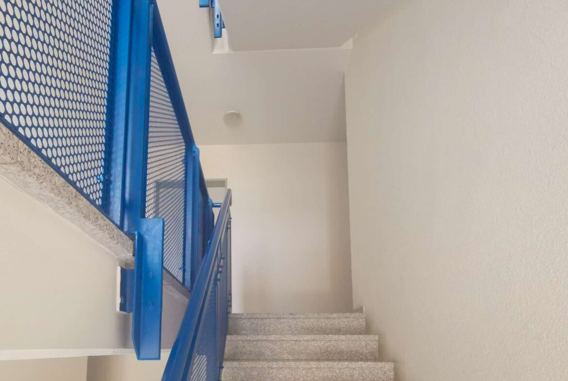 Blue metal stair railing with mesh panels along a stairwell, viewed from the lower steps in a building interior.