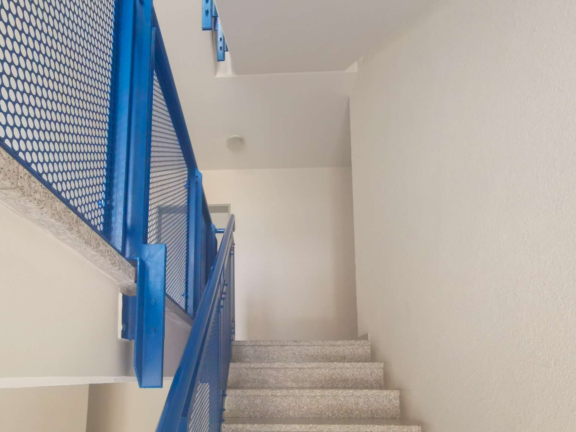 Blue metal stair railing with mesh panels along a stairwell, viewed from the lower steps in a building interior.