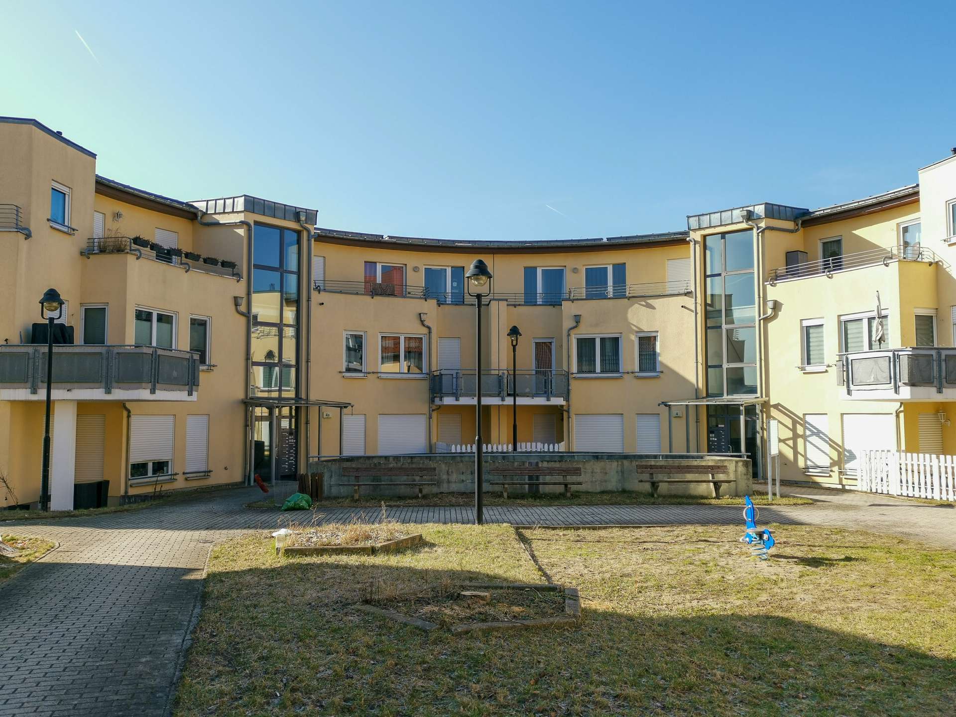 Courtyard of yellow apartment buildings with curved facade, benches, lampposts and a blue spring rider in the foreground against a clear blue sky