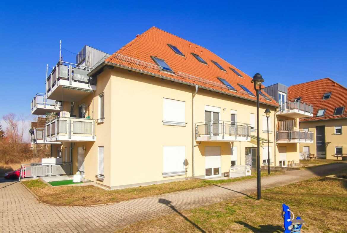 Residential building with orange-tiled roof, balconies, and a blue playground ride in the foreground under a clear blue sky.