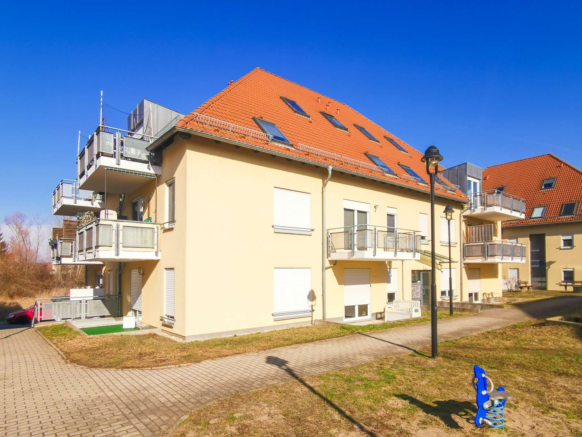Residential building with orange-tiled roof, balconies, and a blue playground ride in the foreground under a clear blue sky.