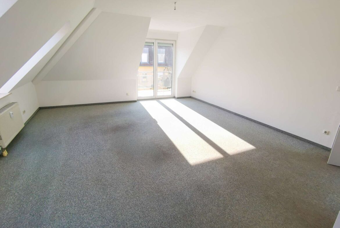 Bright, empty attic room with sloped white walls, a large glass door to a balcony, and sunlight streaming onto a gray carpeted floor.