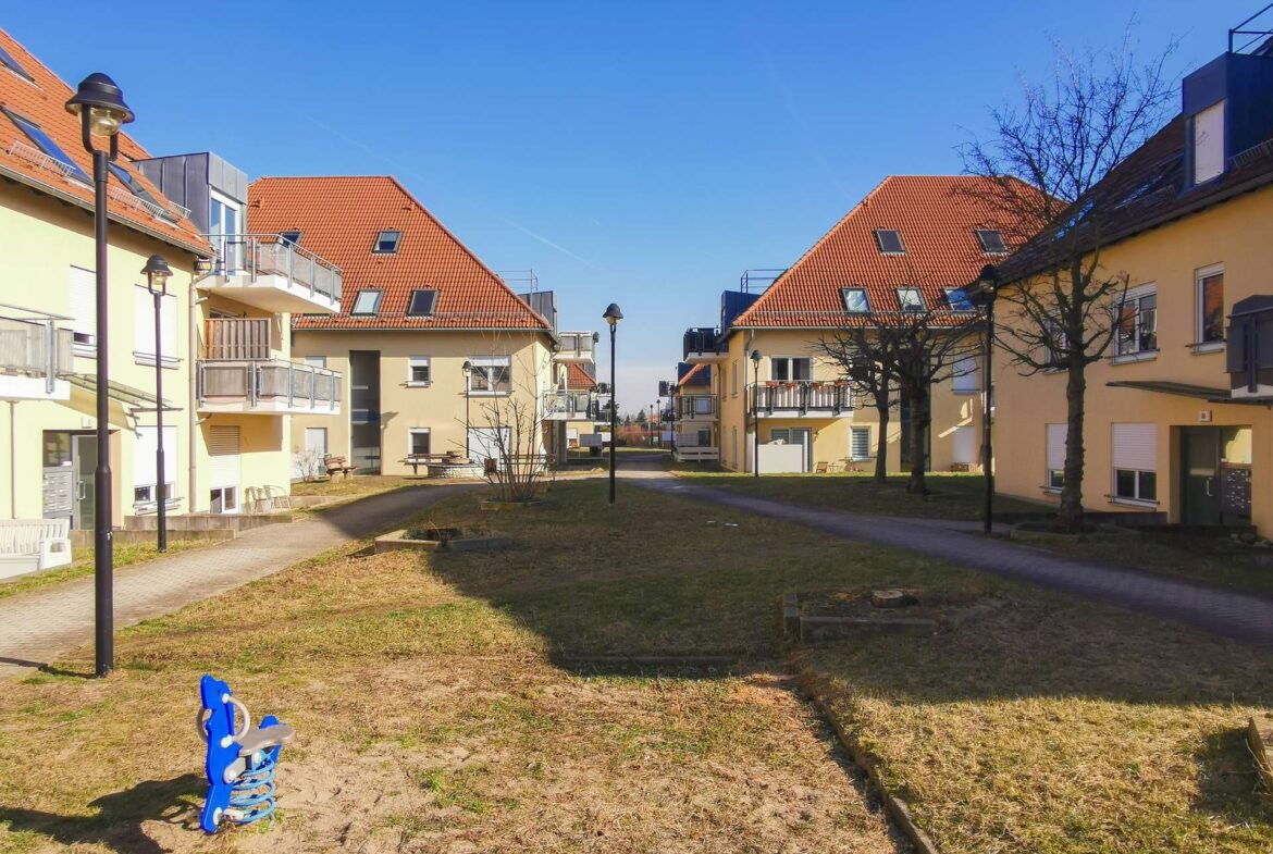 Residential courtyard with yellow apartment buildings, red-tiled roofs, and a blue spring rider in the foreground.