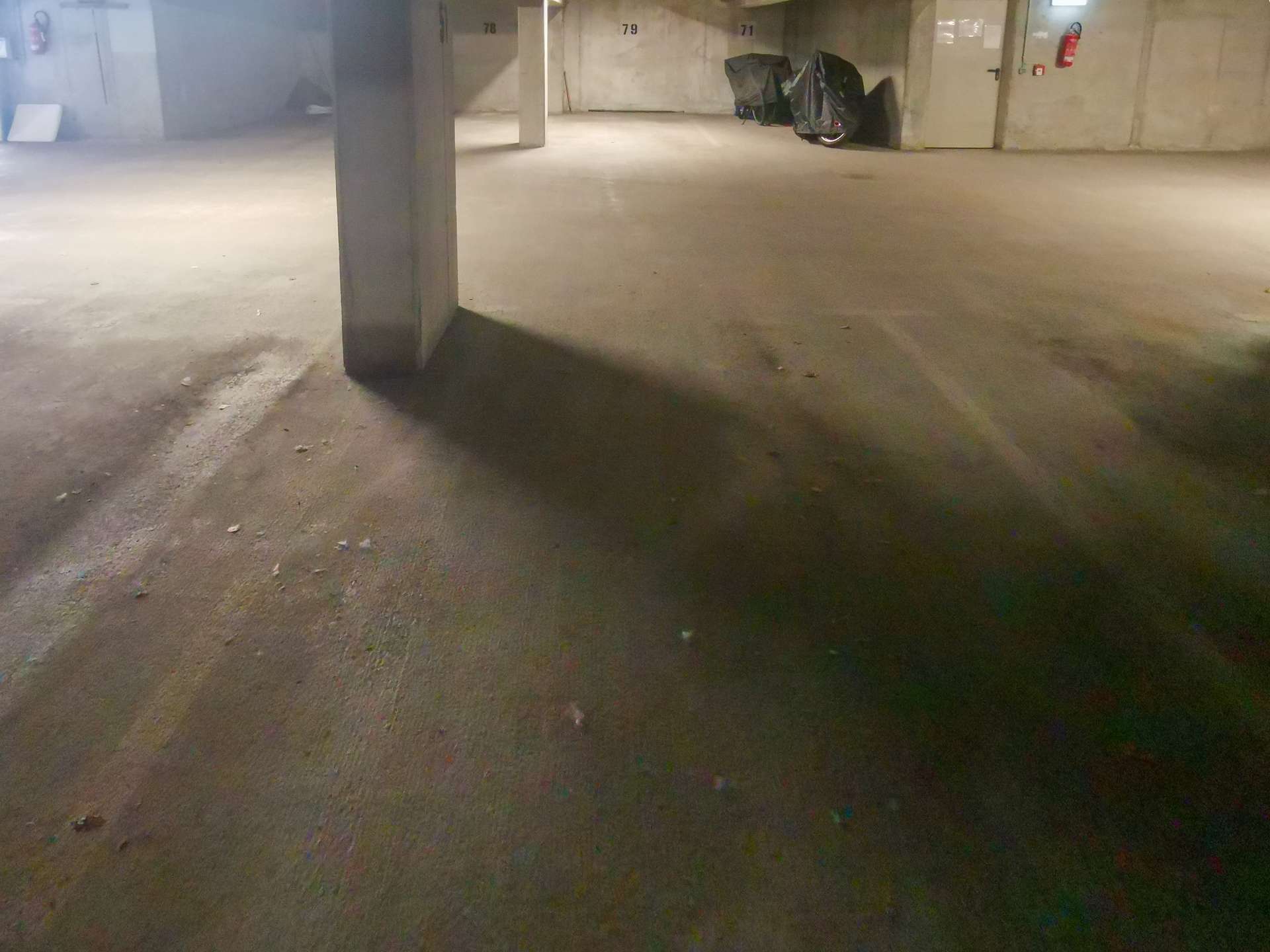Dimly lit underground parking garage with a concrete pillar foreground and a covered scooter near wall 78–79–71 in background on the right side.