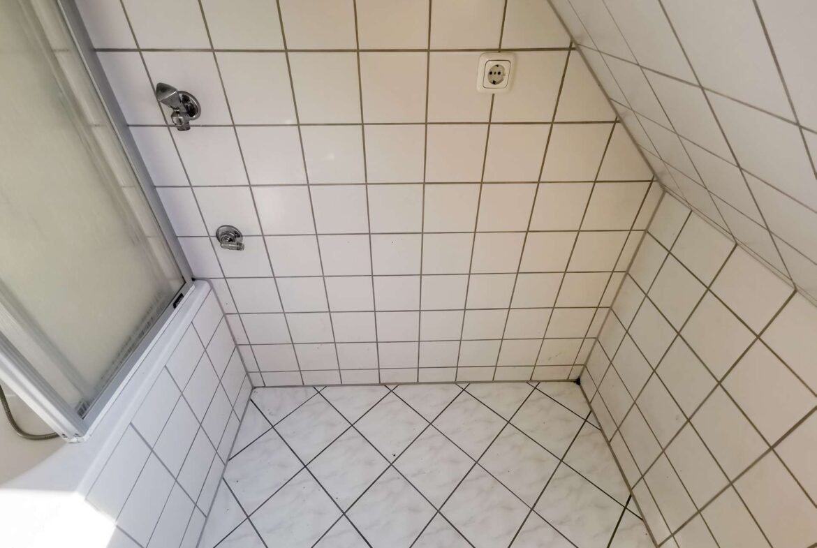 Tile-filled shower corner with a chrome showerhead on the left wall and a white tiled surround, plus a wall outlet near the center.