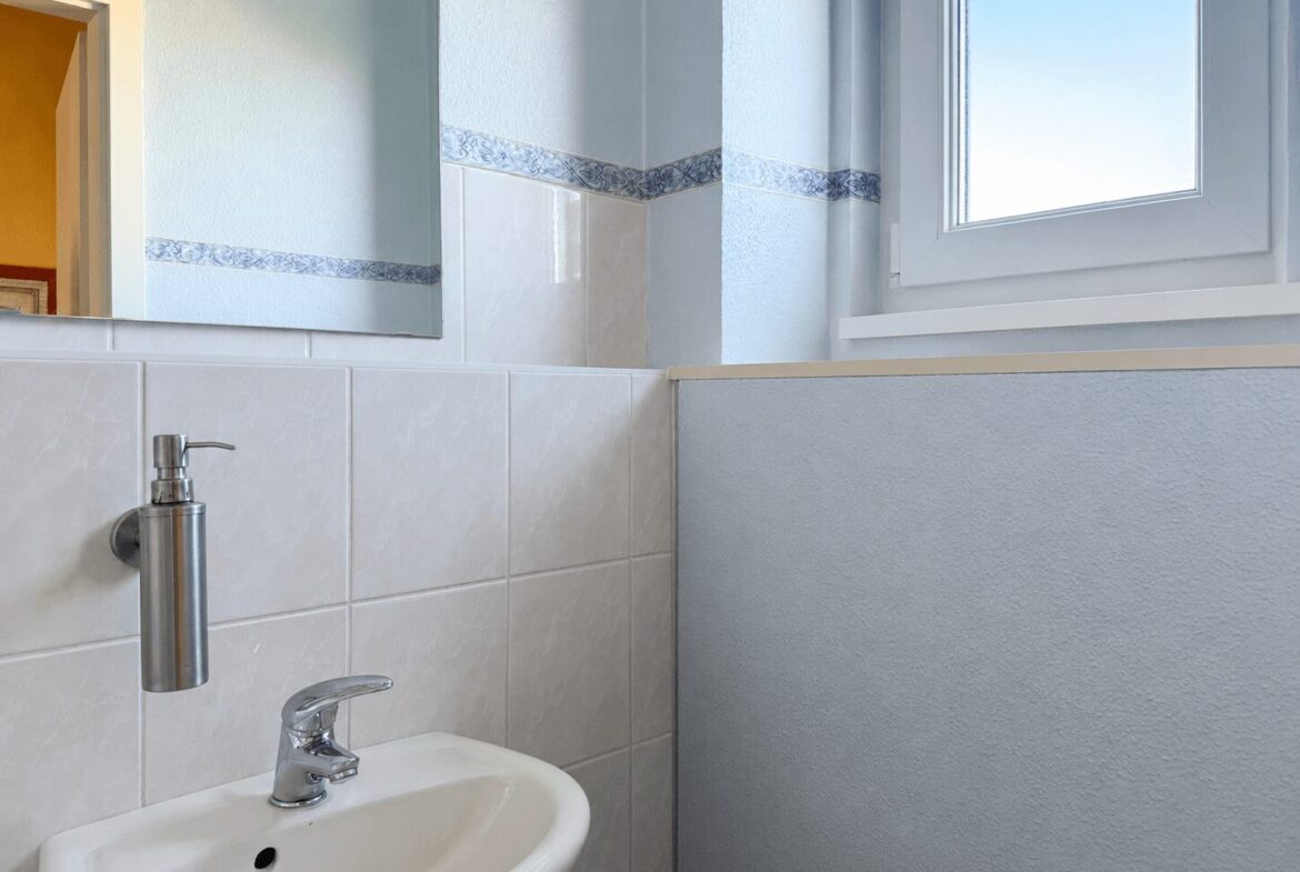 White ceramic sink with a chrome faucet and wall-mounted soap dispenser, in a light blue tiled bathroom with a window on the right.