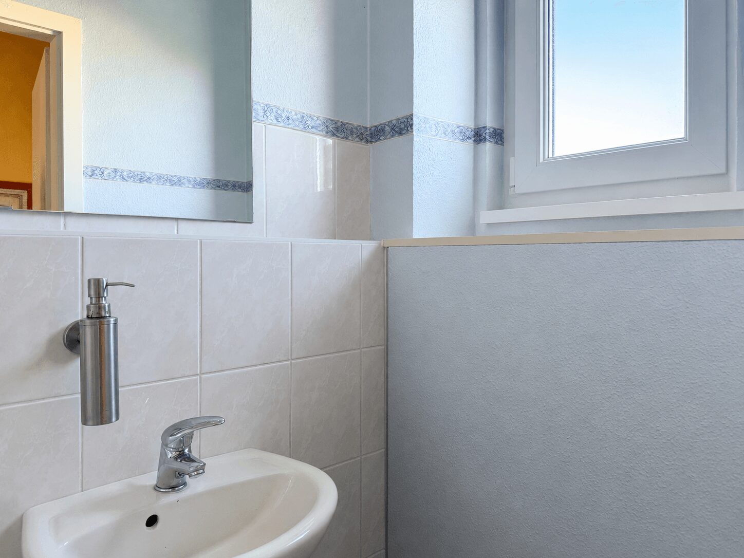 White ceramic sink with a chrome faucet and wall-mounted soap dispenser, in a light blue tiled bathroom with a window on the right.