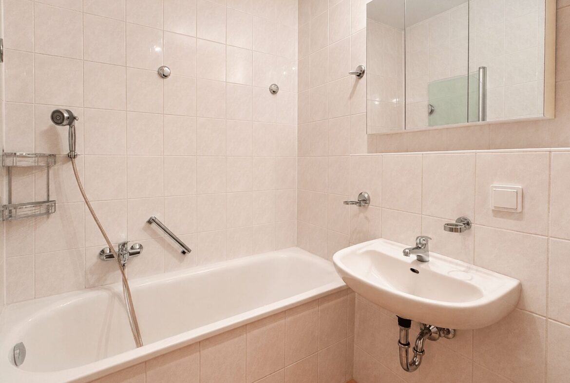 White-tiled bathroom with a bathtub, handheld shower, and a sink with a mirrored cabinet above it.