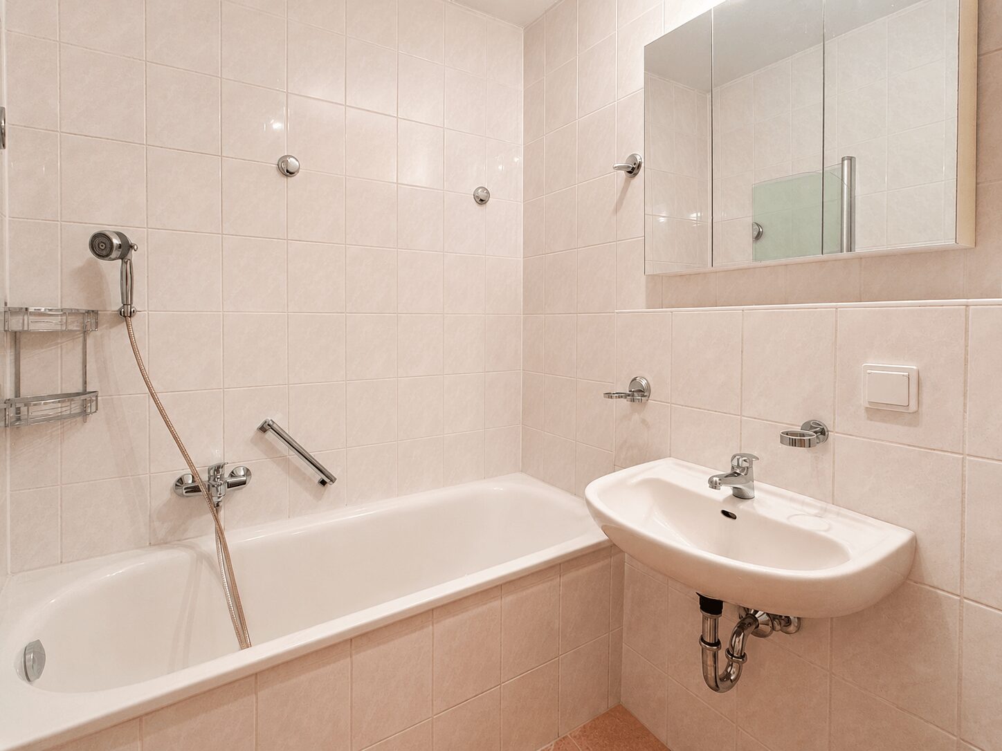 White-tiled bathroom with a bathtub, handheld shower, and a sink with a mirrored cabinet above it.
