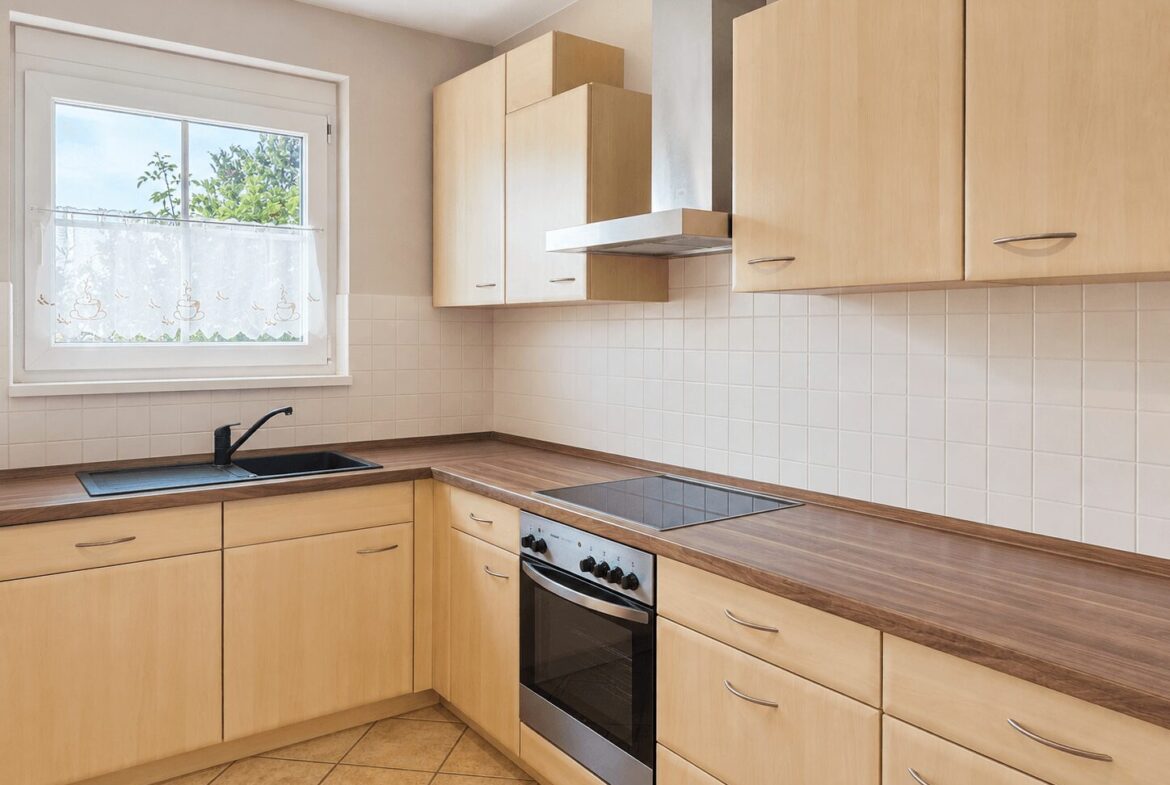 L-shaped kitchen with light wood cabinets, a black sink under a window, and a stainless steel oven with a cooktop and range hood.