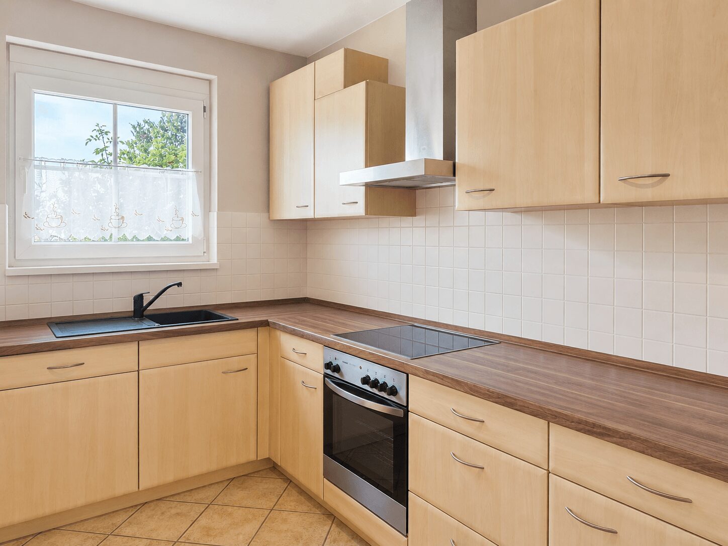 L-shaped kitchen with light wood cabinets, a black sink under a window, and a stainless steel oven with a cooktop and range hood.
