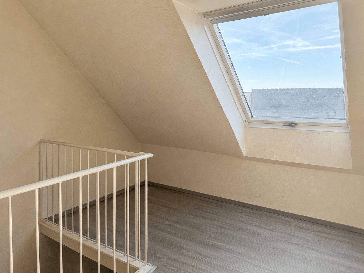 Bright empty attic loft with a slanted ceiling, white railing, and a large skylight letting in daylight.