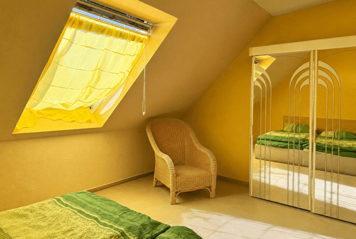 Yellow-toned attic bedroom with a slanted window curtain, wicker chair, and mirrored wardrobe reflecting a green-blanketed bed area.