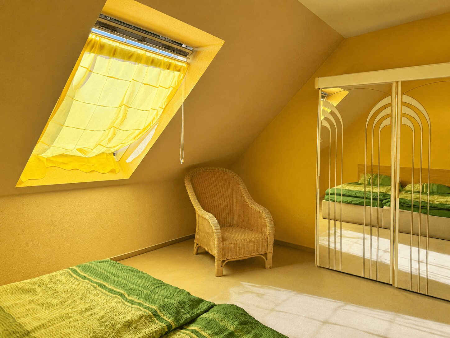 Yellow-toned attic bedroom with a slanted window curtain, wicker chair, and mirrored wardrobe reflecting a green-blanketed bed area.
